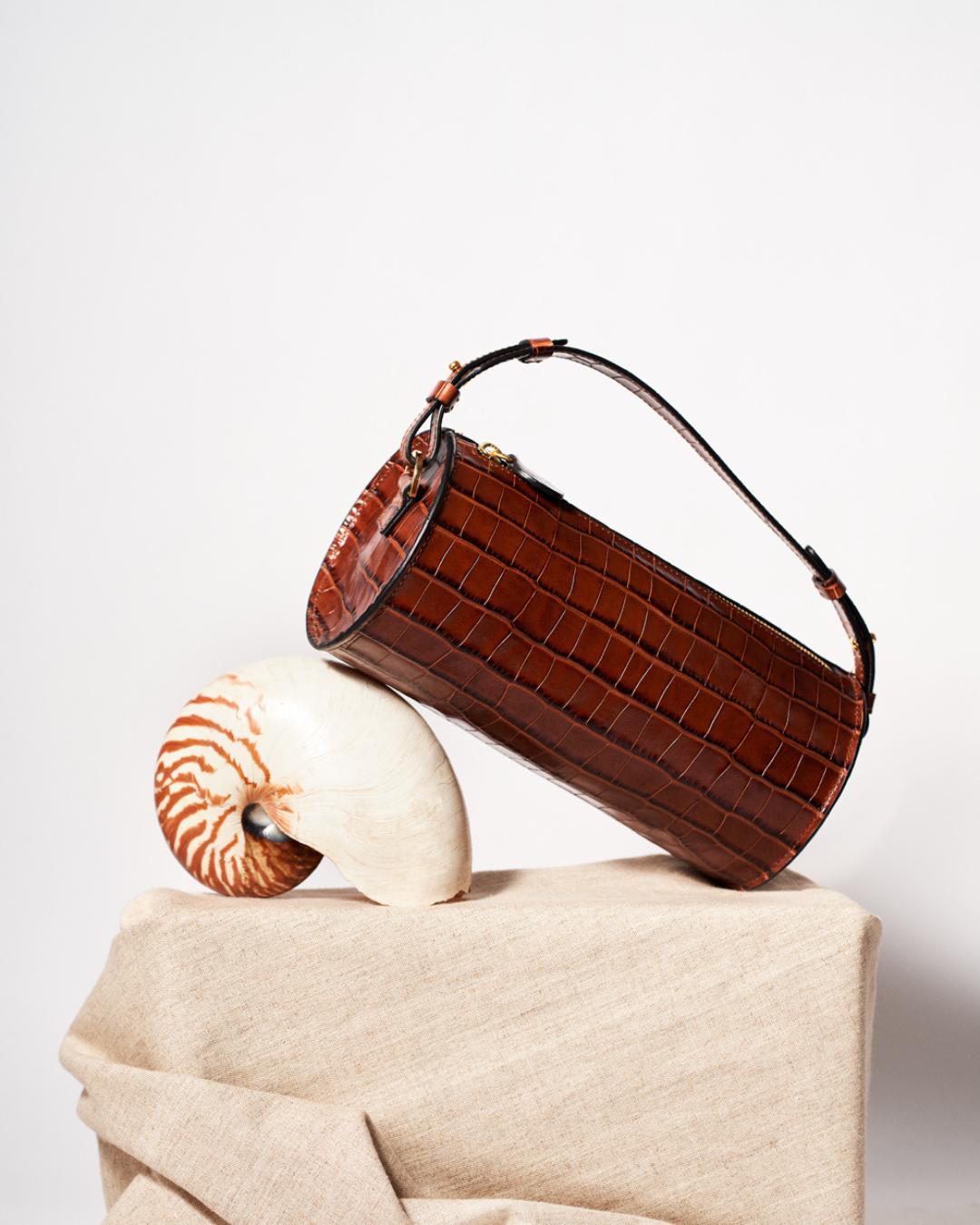 CNicol Brown Croc Leather Evie Bag leaning on White Shell with Brown Stripes on top of a beige fabric