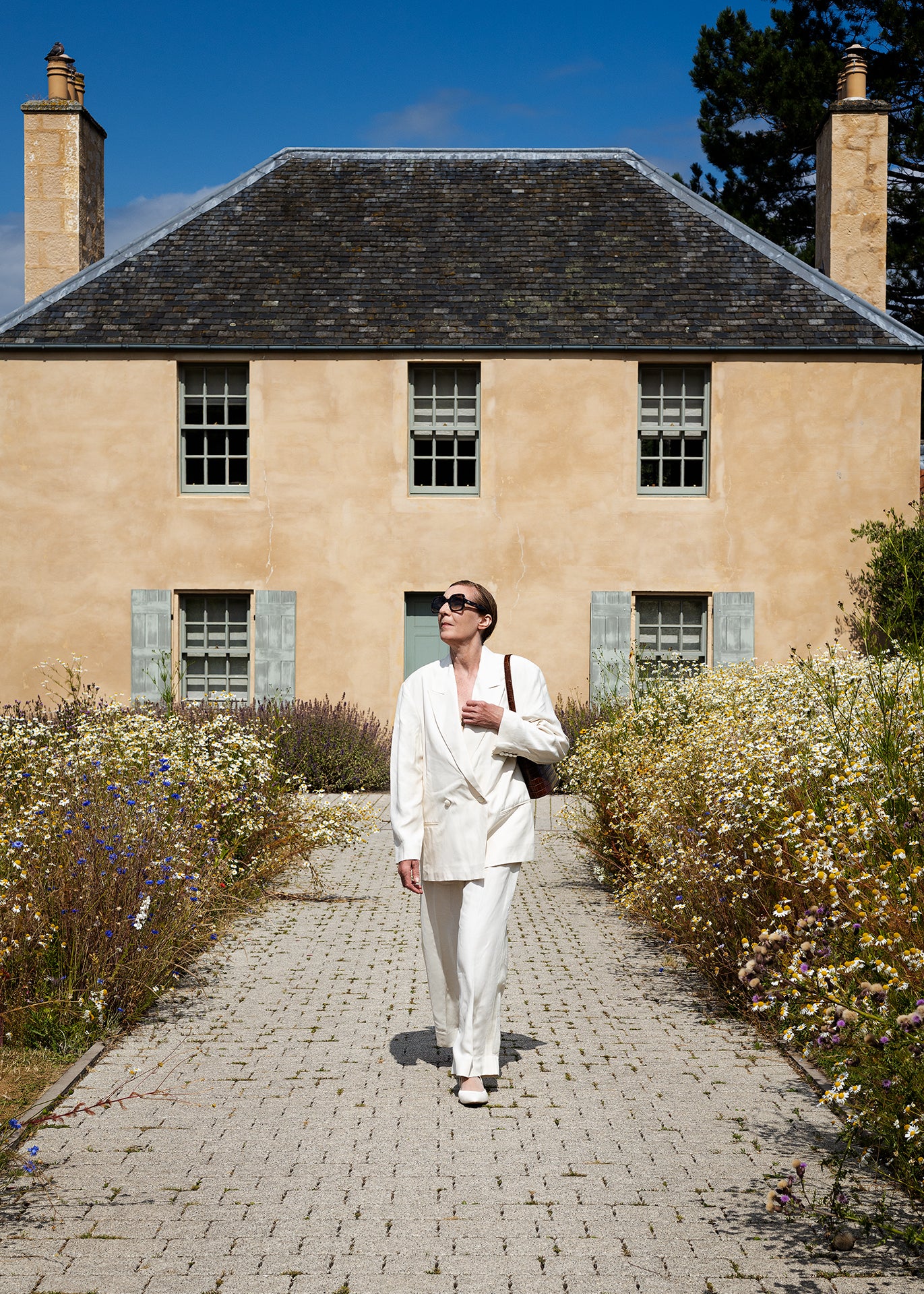 Woman wearing a C.Nicol Scotland brown croco leather bag over her shoulder, while wearing a white suit and walking along a path surrounded by flowers