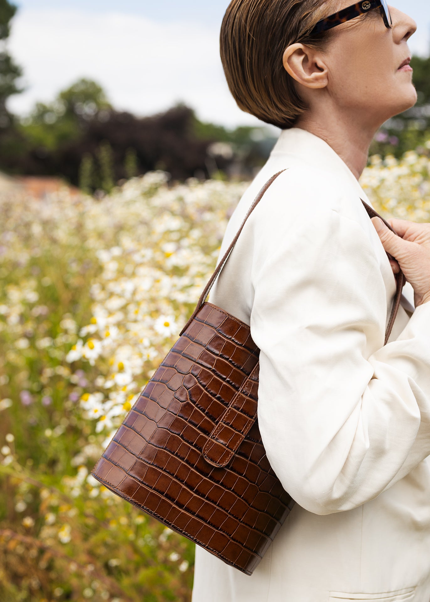 Woman wearing a C.Nicol Scotland brown croco leather bag over her shoulder, while wearing a white suit against a backdrop of flowers 