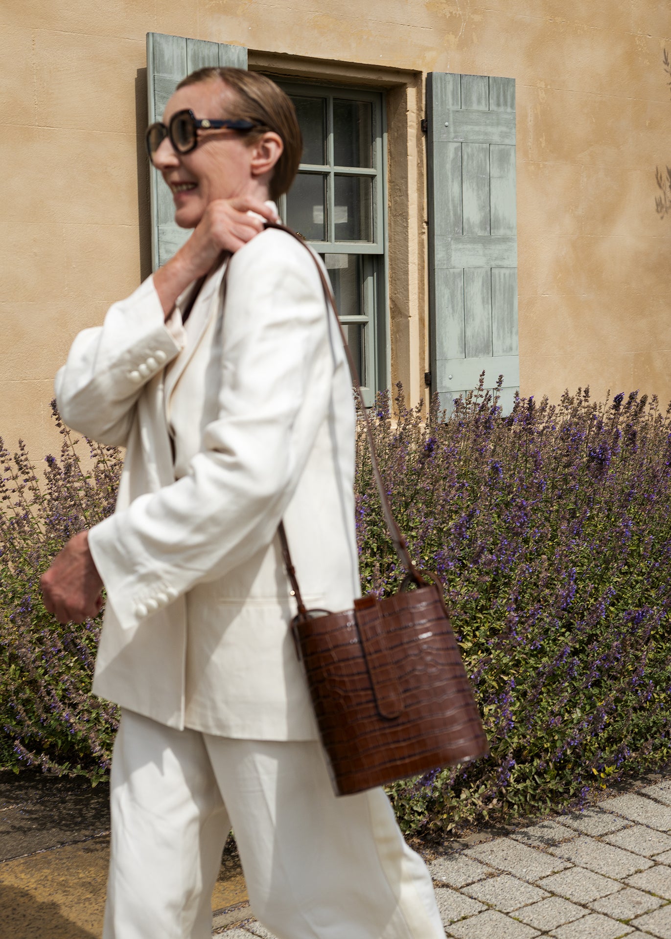 Woman wearing a C.Nicol Scotland brown croco leather bag over her shoulder, while wearing a white suit and walking past a building surrounded with purple flowers