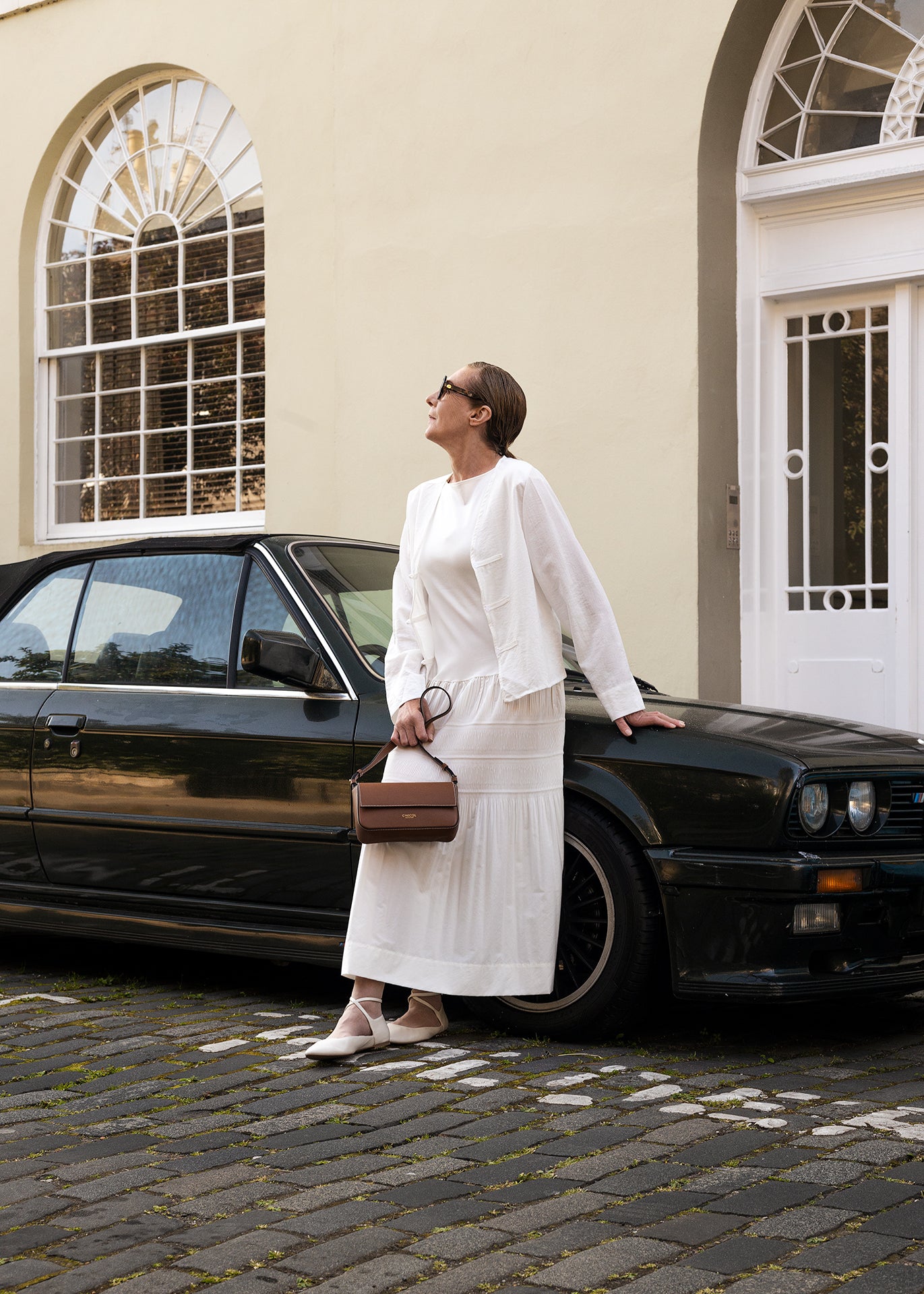 Woman holding a C.Nicol Scotland tan leather bag, wearing a white dress while standing next to a car
