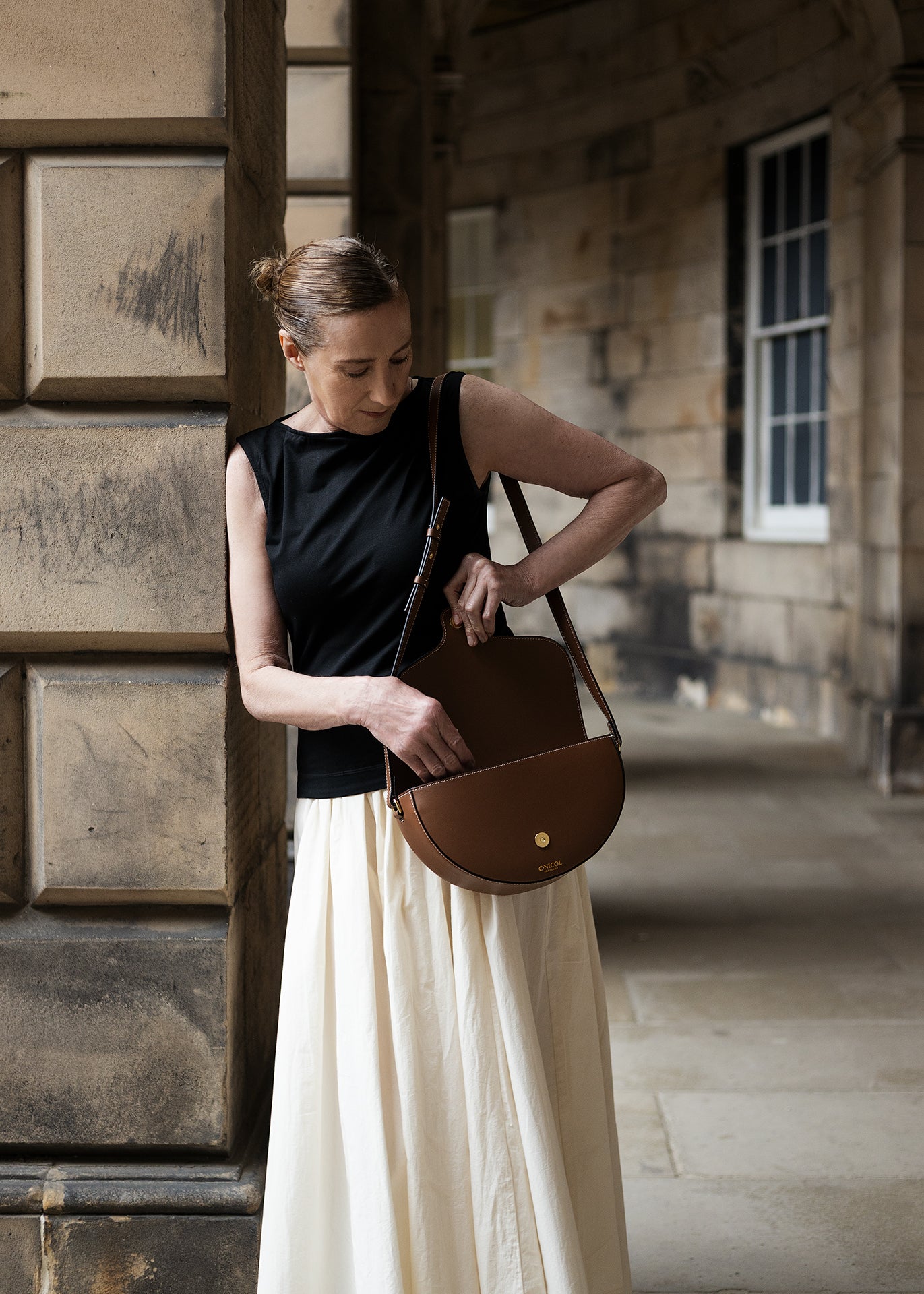 Woman holding  tan leather C.Nicol bag, wearing a black top and white skirt.