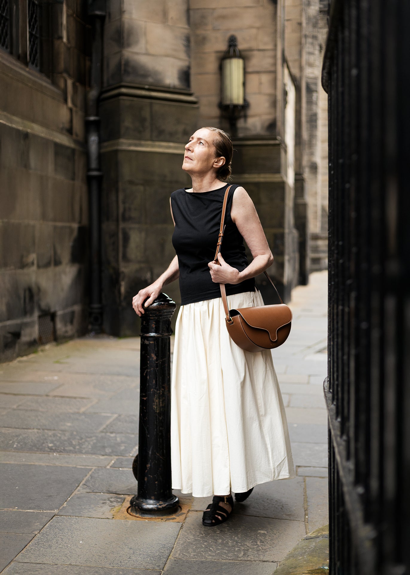 Woman standing on a street corner holding  tan leather C.Nicol bag, wearing a black top and white skirt.